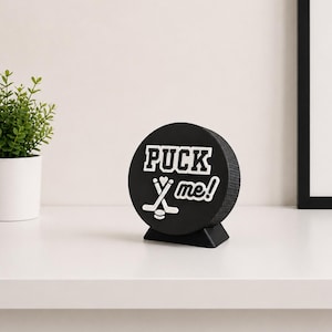 Op de afbeelding: Een zwart decoratief item in de vorm van een hockeypuck met de witte tekst "PUCK ME!". Het ontwerp bevat gekruiste hockeysticks en een puck. Het staat op een kleine zwarte basis.