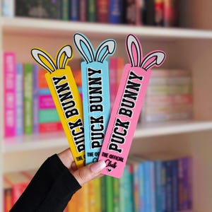 Op de afbeelding: Drie rechthoekige boekenleggers in geel, lichtblauw en roze, elk voorzien van een paar konijnenoren aan de bovenkant. De tekst PUCK BUNNY staat in het zwart op elk exemplaar, met op de roze boekenlegger ook THE OFFICIAL CLUB en een hartje.