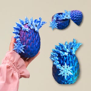 Può includere: Tre sculture di uova di drago fantasy nelle tonalità del blu e del viola. Ogni uovo presenta una testa di drago e dettagli a forma di fiocco di neve. Un uovo è tenuto in mano, mentre gli altri sono esposti separatamente. Le uova hanno un aspetto strutturato e squamoso.
