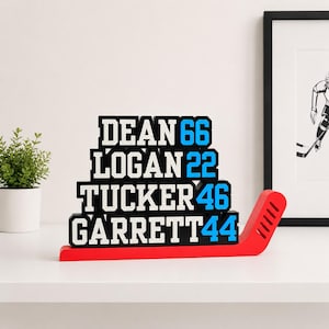 Op de afbeelding: Een hockey-thema naamdisplay in zwart, wit en blauw. De namen DEAN, LOGAN, TUCKER en GARRETT zijn gestapeld met hun rugnummers. Het rust op een rode hockeystick basis.