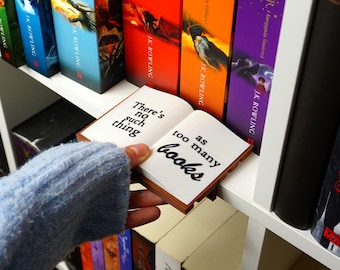 Könnte beinhalten: Ein kleiner, offener Lesezeichen in Buchform mit dem Text "There's no such thing as too many books" in Schwarz. Das Lesezeichen wird in der Nähe eines Regals mit farbigen Büchern gehalten, darunter Titel von J.K. Rowling.