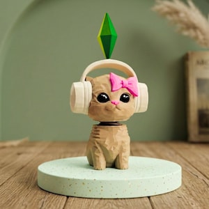 Pode incluir: Uma estatueta de gato castanho com grandes olhos pretos, usando fones de ouvido brancos e um laço rosa. Uma forma de diamante verde e amarelo está acima da cabeça do gato. O gato está em uma plataforma verde clara salpicada.