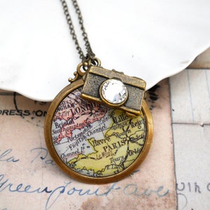 May include: A vintage-style necklace featuring a round pendant with a map design and a small camera charm. The map pendant shows a section of Europe with cities like London and Paris. The camera charm has a clear crystal. The necklace has a bronze finish.