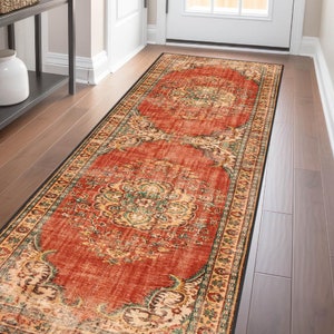 Può includere: Un lungo tappeto rettangolare con un design ornato e usurato. Il tappeto presenta uno sfondo prevalentemente rosso con motivi intricati nelle tonalità del crema, verde e oro. Un bordo nero incornicia il tappeto, posto su un pavimento in legno.