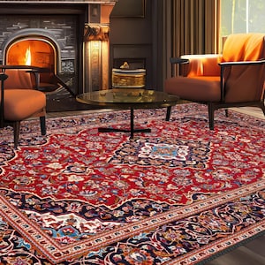 May include: A detailed shot of a living room featuring a large, ornate red rug with a floral pattern. Two orange armchairs face a fireplace with a burning fire. A round coffee table sits in the center, and a painting hangs above the fireplace.
