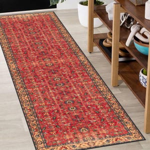 May include: A long rectangular runner rug featuring a vibrant red base colour with an intricate floral and vine pattern in shades of blue, green, and gold. The rug is framed by a decorative border with a repeating motif in tan and dark blue tones.