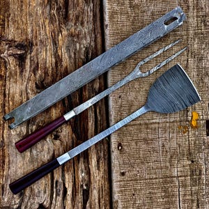 Puede incluir: Un juego de tres utensilios de cocina con mangos de madera. Los utensilios están hechos de acero inoxidable con un acabado martillado. El juego incluye una espátula, un tenedor y una herramienta larga y delgada.