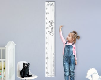 May include: White wooden growth chart with a distressed wood finish. The chart features a ruler with measurements in inches and feet. The text "loved beyond measure" is at the top of the chart. The name "Charlotte" is written in cursive on the right side of the chart. A young girl is standing next to the chart, measuring her height.