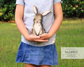 May include: A child is holding a brown and white rabbit cutout with a red heart on its chest. The rabbit is sitting on its hind legs with its front paws together. The child is wearing a gray t-shirt and a blue denim skirt. The background is a blurry green lawn.