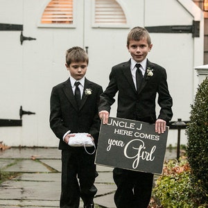 May include: Two young boys in black suits stand in front of a white door. The boy on the right holds a sign that says "Uncle J Here Comes Your Girl".