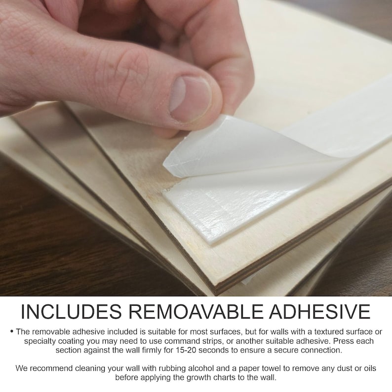 May include: Close-up of a white removable adhesive strip attached to a wooden surface. The adhesive is designed for use on most surfaces, including walls. The text "INCLUDES REMOVABLE ADHESIVE" is visible at the bottom of the image.