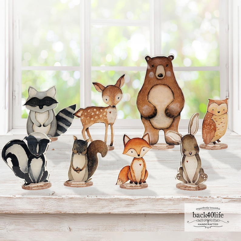 May include: A set of nine wooden animal figurines, including a bear, deer, raccoon, owl, fox, rabbit, and two squirrels. The figurines are painted in realistic colours and have a whimsical, hand-painted style. The figurines are standing on a white wooden surface.