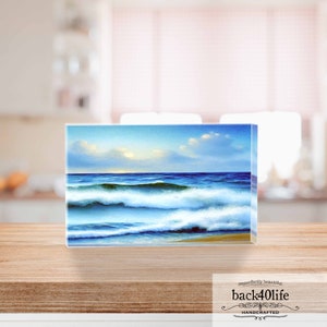 May include: A painting of a seascape with blue and white waves crashing on a sandy beach. The sky is a light blue with white clouds. The painting is framed in a clear acrylic block.