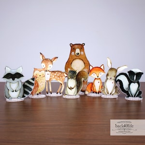 May include: A set of eight woodland animal figurines, including a raccoon, owl, deer, bear, squirrel, fox, rabbit, and skunk. The figurines are painted in a watercolour style with brown, grey, white, and black colours. The figurines are standing on clear acrylic bases.