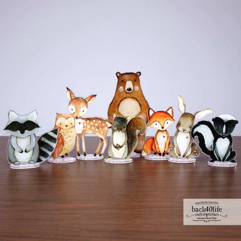 May include: A set of eight woodland animal figurines, including a raccoon, owl, deer, bear, squirrel, fox, rabbit, and skunk. The figurines are painted in a watercolour style with brown, grey, white, and black colours. The figurines are standing on clear acrylic bases.