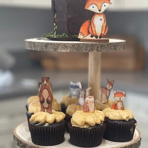 May include: A two-tiered wooden cake stand with a brown chocolate cake on the top tier. The bottom tier has six cupcakes decorated with brown, tan, and orange animal toppers. The toppers include a bear, an owl, a fox, a raccoon, and a deer.
