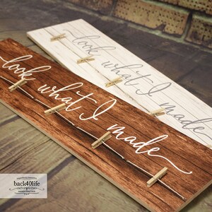 May include: A wooden sign with the text "look what I made" written in white cursive script. The sign has a rustic brown wood finish and is decorated with small wooden clothespins and twine. The sign is perfect for displaying photos or other small items.