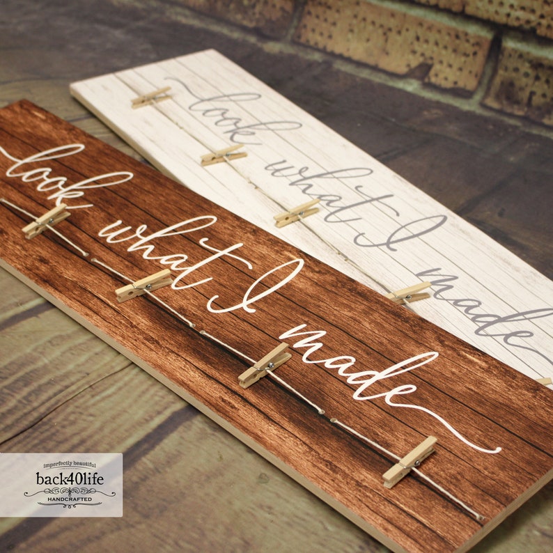 May include: A wooden sign with the text "look what I made" written in white cursive script. The sign has a rustic brown wood finish and is decorated with small wooden clothespins and twine. The sign is perfect for displaying photos or other small items.
