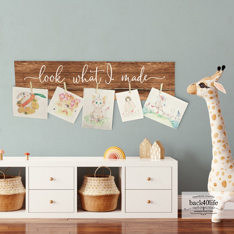 May include: A wooden sign with the text "look what I made" hanging on a light blue wall.  The sign has six clothespins holding colorful drawings of animals and people.  Below the sign is a white shelf with three drawers and a woven basket.  A stuffed giraffe stands on the right side of the image.