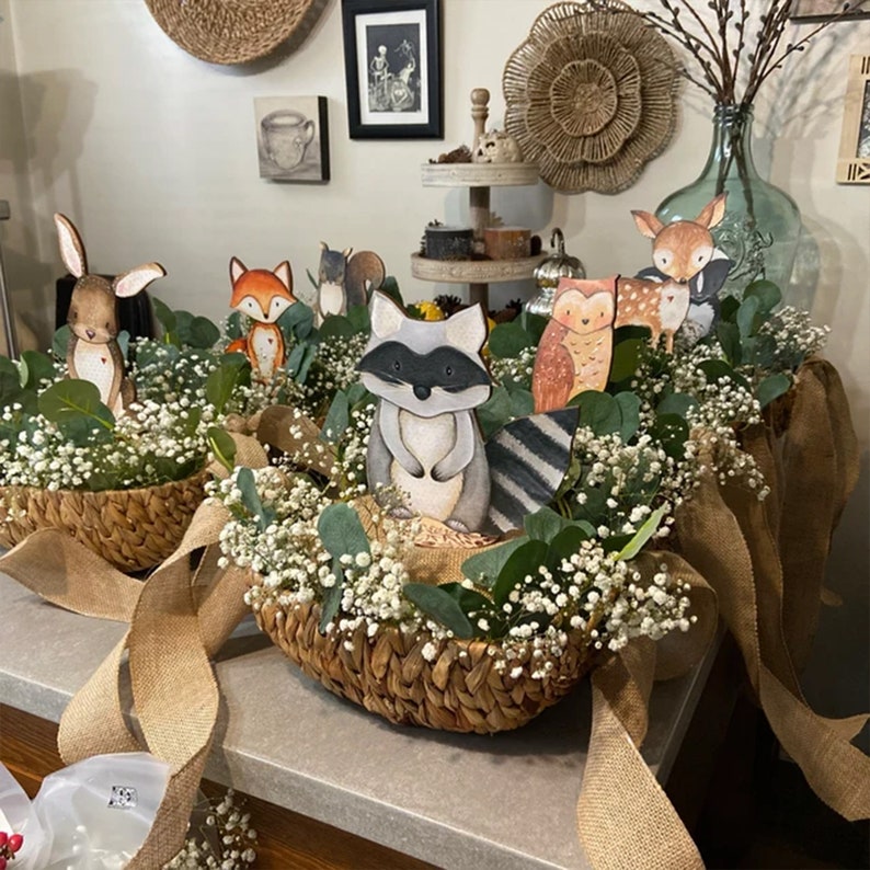 May include: A woodland themed centrepiece featuring a variety of animal figurines, including a raccoon, fox, owl, deer, and rabbit. The figurines are arranged in a woven basket with white baby's breath and greenery.