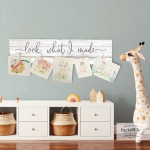 May include: White wooden sign with the text "look what I made" hanging on a wall with six colorful drawings of animals and people hanging from clothespins. A white bookshelf with two drawers and a woven basket sits below the sign. A stuffed giraffe stands to the right of the sign.