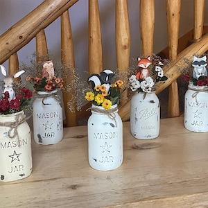May include: Five white mason jars with different animal figurines on top. The jars are decorated with black paint and the words "Mason Jar" are written on them. The jars are filled with flowers and greenery.
