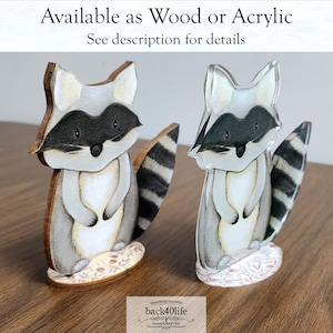 May include: Two raccoon figurines, one made of wood and one made of acrylic. Both figurines are painted in a watercolour style with a white background and black and grey stripes. The figurines are standing on a base with a floral design. The text "Available as Wood or Acrylic" and "See description for details" is at the top of the image. The text "imperfectly beautiful" and "back40life HANDCRAFTED" is at the bottom of the image.