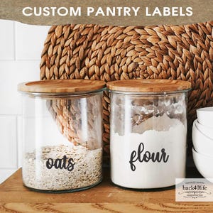 May include: Two glass jars with wooden lids, filled with oats and flour. The jars have black handwritten labels that say "oats" and "flour".
