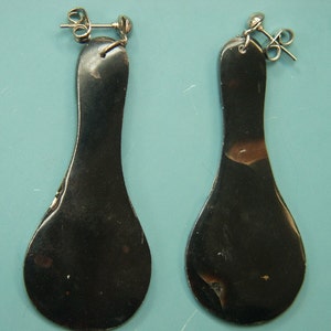 May include: A pair of dark, elongated earrings with a teardrop shape. The earrings have a shiny, metallic appearance with a silver post and backing. The earrings are displayed against a light blue background.