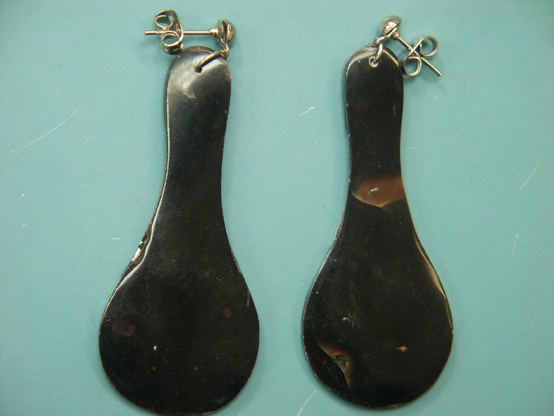 May include: A pair of dark, elongated earrings with a teardrop shape. The earrings have a shiny, metallic appearance with a silver post and backing. The earrings are displayed against a light blue background.