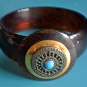 Op de afbeelding: Een bruine plastic armband met een ronde, goudkleurige, zwart-zilveren accent. De accent heeft een turquoise blauwe steen in het midden.