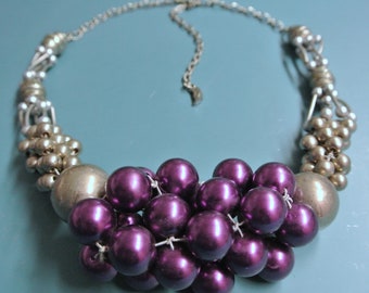 May include: A statement necklace featuring a cluster of deep purple, round beads, resembling grapes, suspended from a silver-toned chain. The necklace has silver and gold-toned accents, creating a bold and eye-catching design.