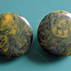 Puede incluir: Un par de pendientes redondos de color negro y amarillo con vetas. Los pendientes tienen un patrón de remolino.