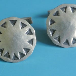May include: A pair of silver cufflinks with a circular design. Each cufflink features a series of triangular shapes around the edge. The cufflinks are set against a light blue background.