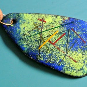 May include: Abstract pendant with a blue, green, and yellow speckled design. The pendant has a brown leather cord and a gold-colored metal loop. The pendant features colorful lines in red, yellow, green, and gray.