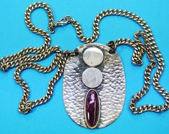 May include: A silver oval pendant necklace with a hammered texture. The pendant features three silver circles and a purple oval stone.