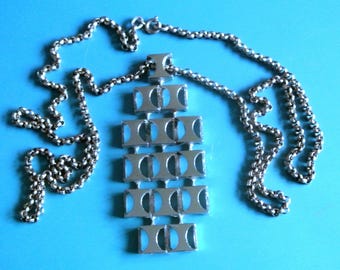 Puede incluir: Collar plateado con colgante geométrico. El colgante presenta una serie de formas rectangulares con cortes redondeados, suspendidas de una cadena. El collar se muestra sobre un fondo azul, resaltando su brillo metálico.