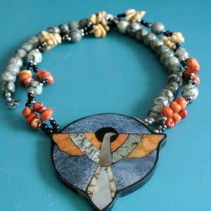 May include: A handcrafted necklace with a bird pendant in orange, grey, and white. The necklace has multiple strands of beads in grey, orange, black, and beige. This unique necklace is a piece of jewellery.