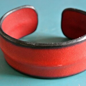 May include: A red leather bracelet with a black trim. The bracelet has a simple, elegant design.