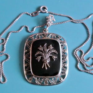 May include: A silver necklace with a black enamel pendant featuring a stylized floral design. The pendant is surrounded by a silver filigree border.