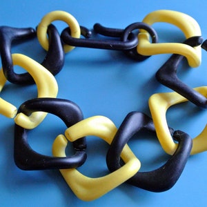 Puede incluir: Un collar de cadena de plástico negro y amarillo con un diseño geométrico. El collar está hecho de eslabones entrelazados que tienen forma de cuadrados y triángulos.