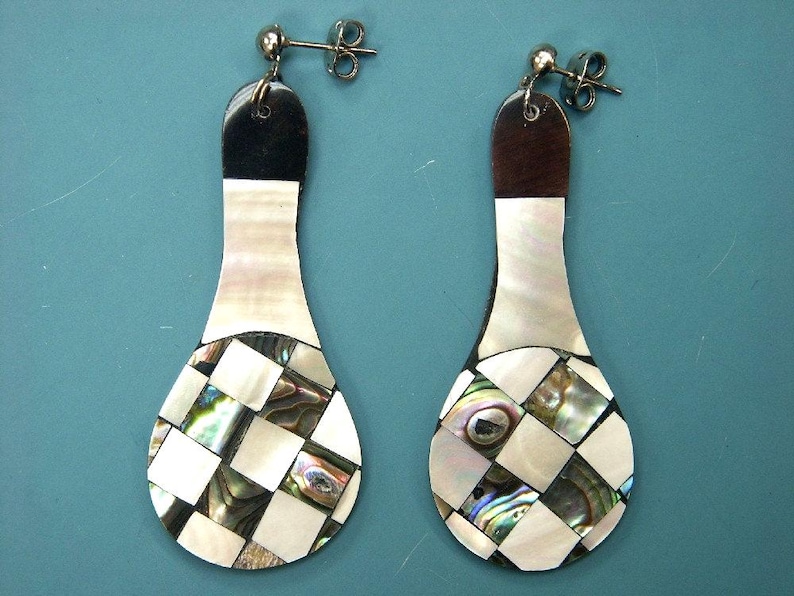 May include: A pair of drop earrings with a distinctive design. The earrings have a black top, a white and iridescent shell middle section, and a checkered pattern at the bottom. They have silver-coloured ear wires.