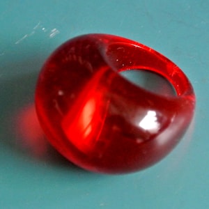 May include: A vibrant, translucent red ring with a rounded, organic shape. The ring is made of a glossy material, and the light reflects off its smooth surface. The ring is a bold statement piece, perfect for adding a pop of color to any outfit.