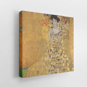 Puede incluir: Impresión en lienzo de una pintura de Gustav Klimt, que representa a una mujer con un vestido dorado con patrones intrincados. La obra de arte tiene un fondo dorado con formas geométricas y toques de rojo y azul.