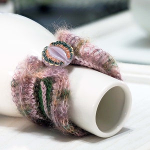 May include: A pink, green, and brown crocheted bracelet with a decorative button featuring a light purple oval center and gold accents.