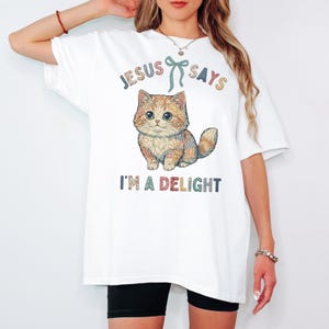 May include: White t-shirt featuring a cartoon cat with the text "JESUS SAYS I'M A DELIGHT". The cat is orange and brown with blue eyes. The text is in a colorful, playful font.