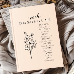 Puede incluir: Un diario rosa con el nombre "sarah" escrito en la portada. El diario tiene un diseño floral y el texto "Dios dice que eres" con una lista de afirmaciones positivas y versículos bíblicos.