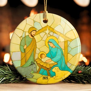 May include: Round Christmas ornament featuring a stained-glass style depiction of the Nativity scene. The ornament shows Mary, Joseph, and baby Jesus, with a donkey and a lamb. The year "2025" is printed on the bottom.