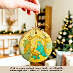 May include: Round Christmas ornament with a stained-glass style nativity scene. The ornament features Joseph, Mary, and baby Jesus, with a donkey and sheep. The year 2025 is printed on the ornament. The ornament is held by a gold string.