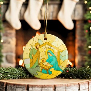 May include: A round Christmas ornament featuring a Nativity scene. The ornament depicts Mary, Joseph, and baby Jesus in a manger, with a donkey and a lamb. The design is in a stained-glass style, with a gold hanging string.
